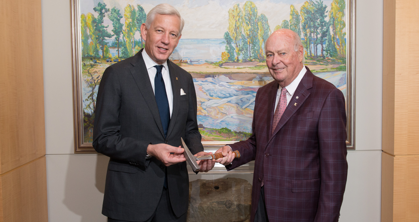 Two men in suits standing in front of a painting, one handing the other a small object.
